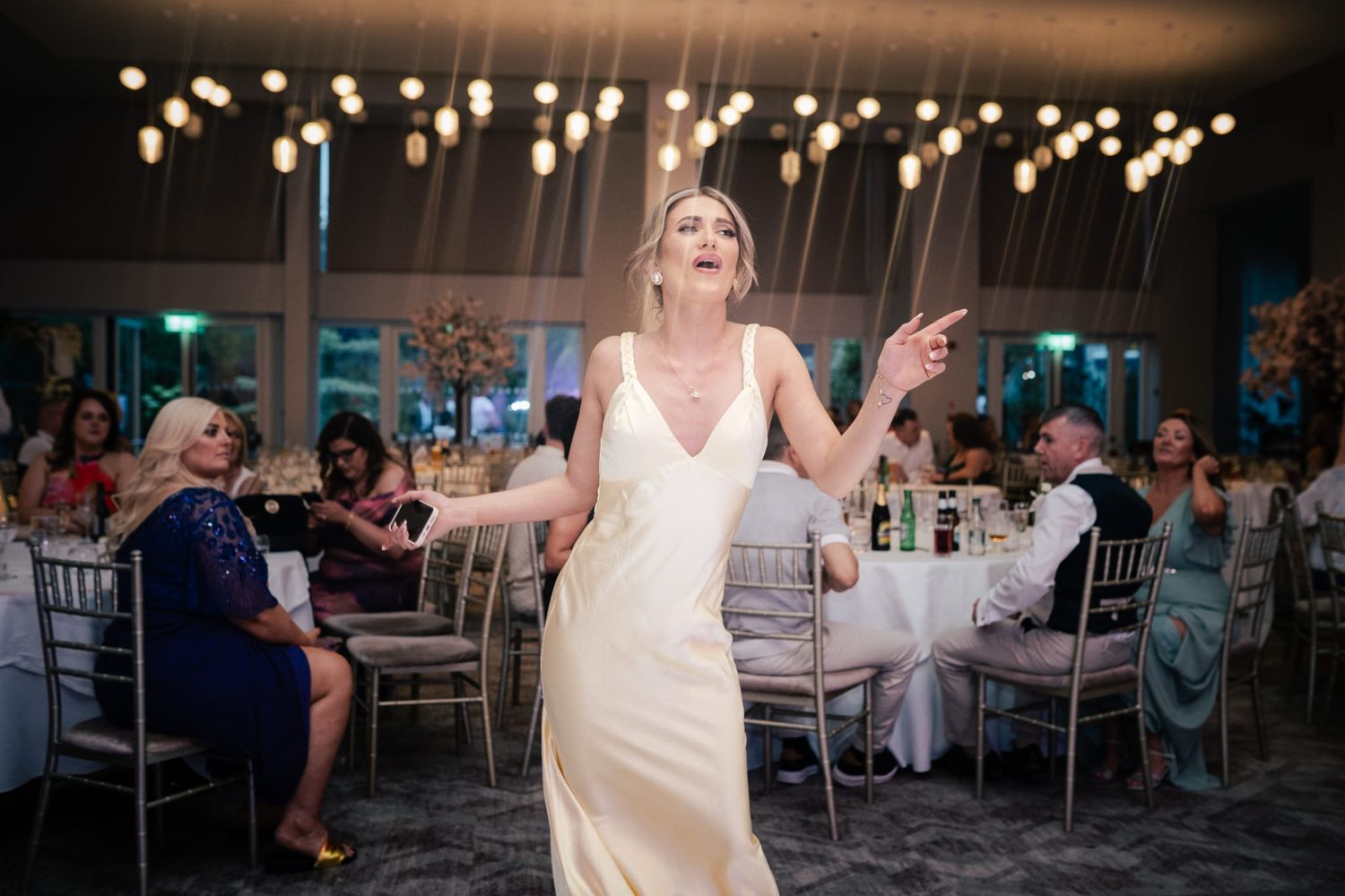 A joyful person in a white wedding dress dances at a reception venue decorated with twinkling lights and elegant tables.