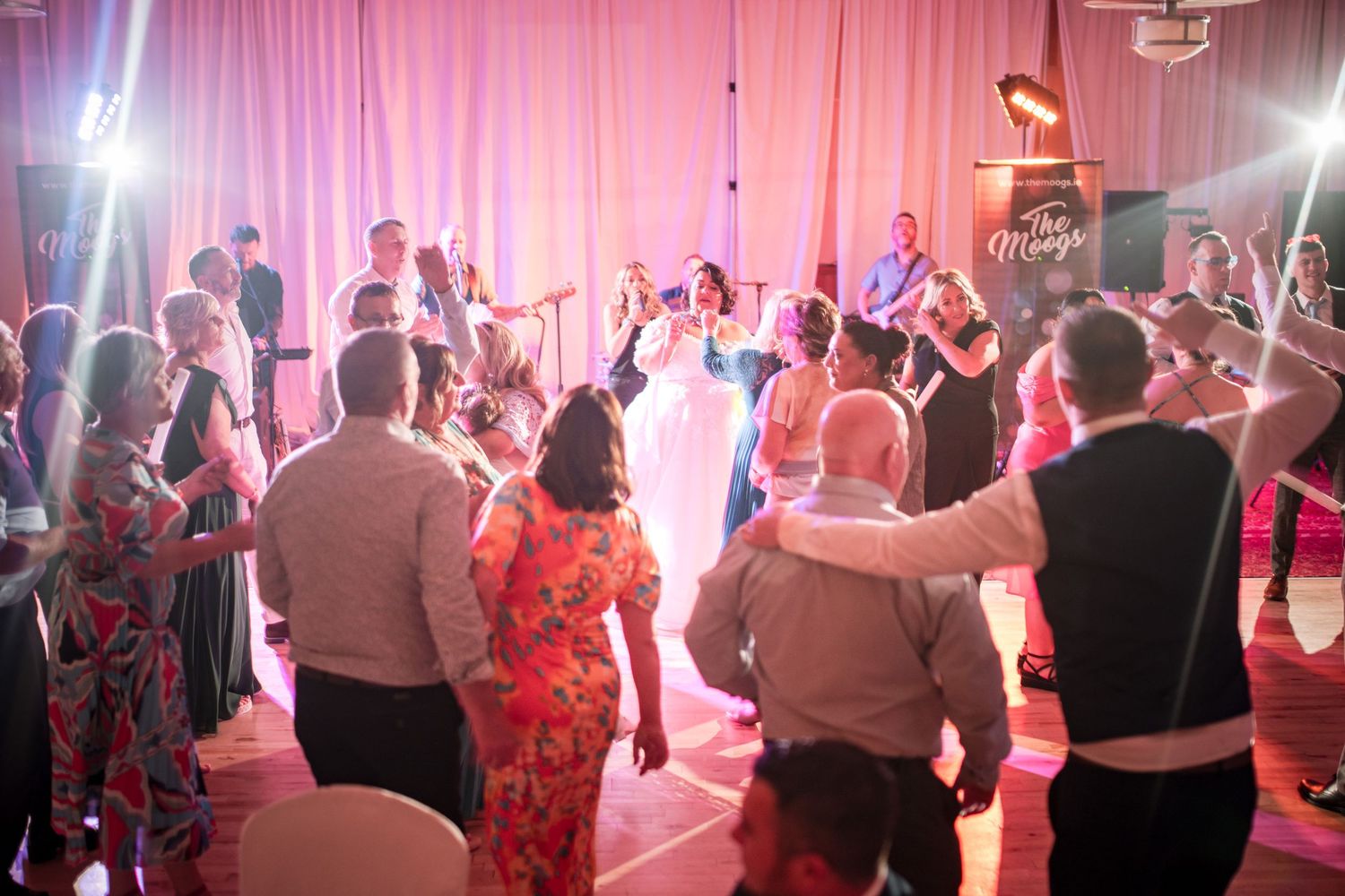 Wedding reception dance floor filled with guests dancing under pink lighting and bright spotlights.