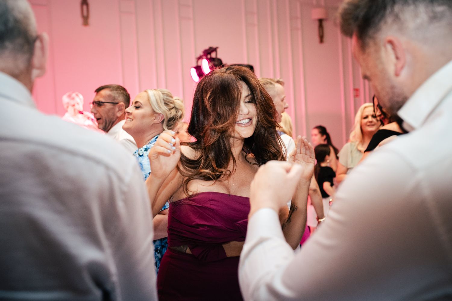 A group dances and celebrates together at a wedding reception with pink lighting in the background.
