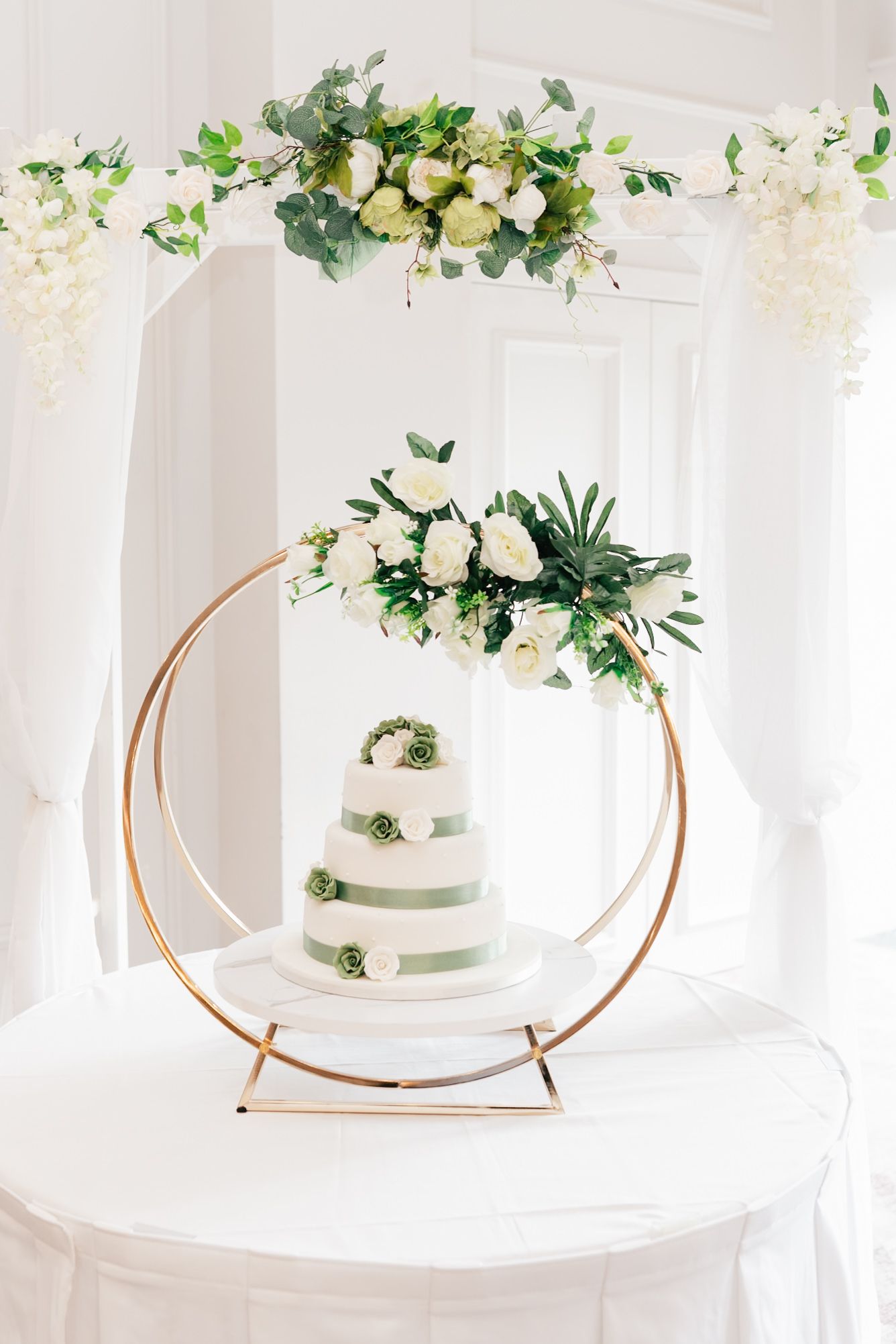 Three elegant white wedding cakes displayed on gold circular stands with cascading white and green floral decorations.