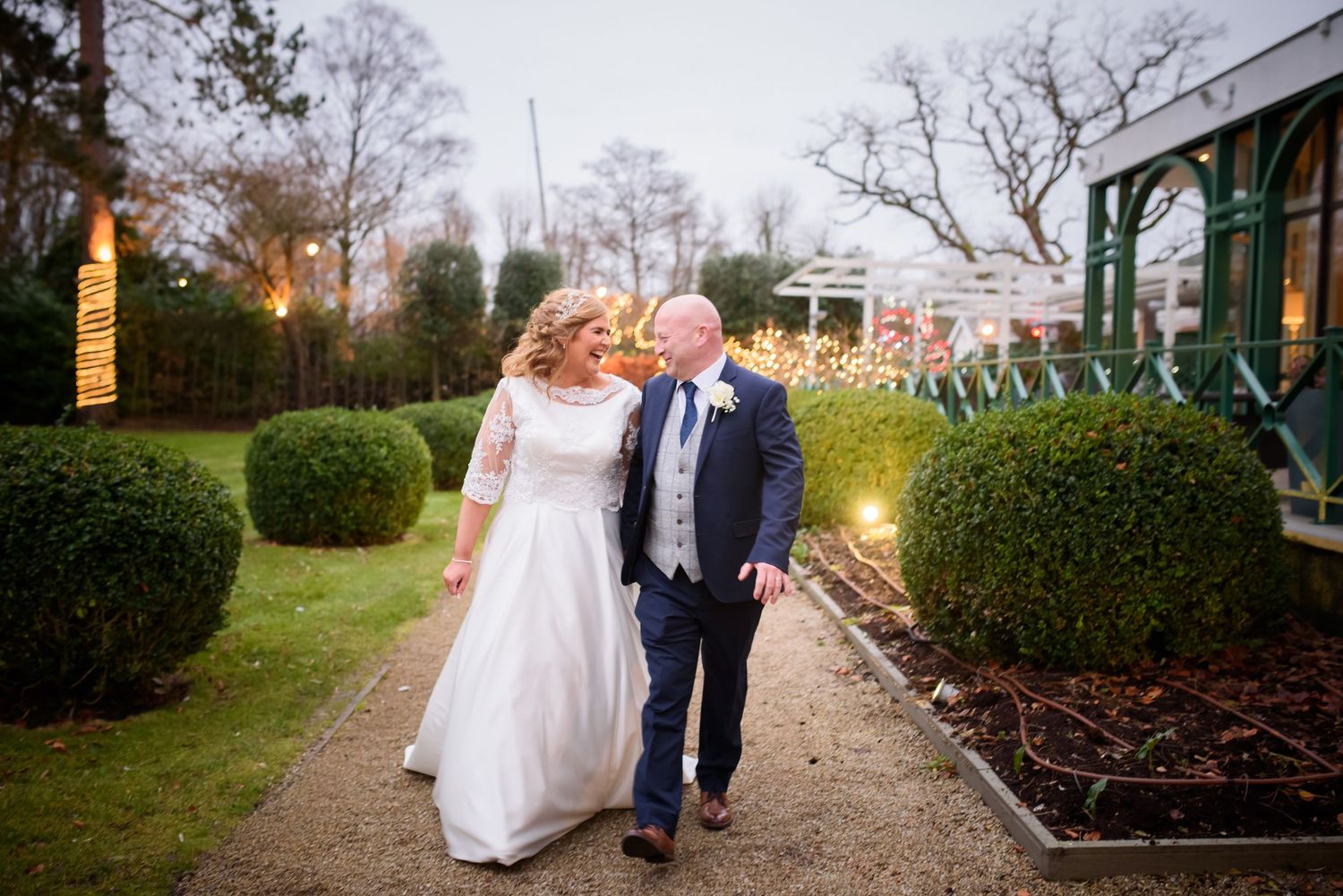Two people in formal attire walk down a garden path lined with manicured hedges at a wedding venue at dusk.