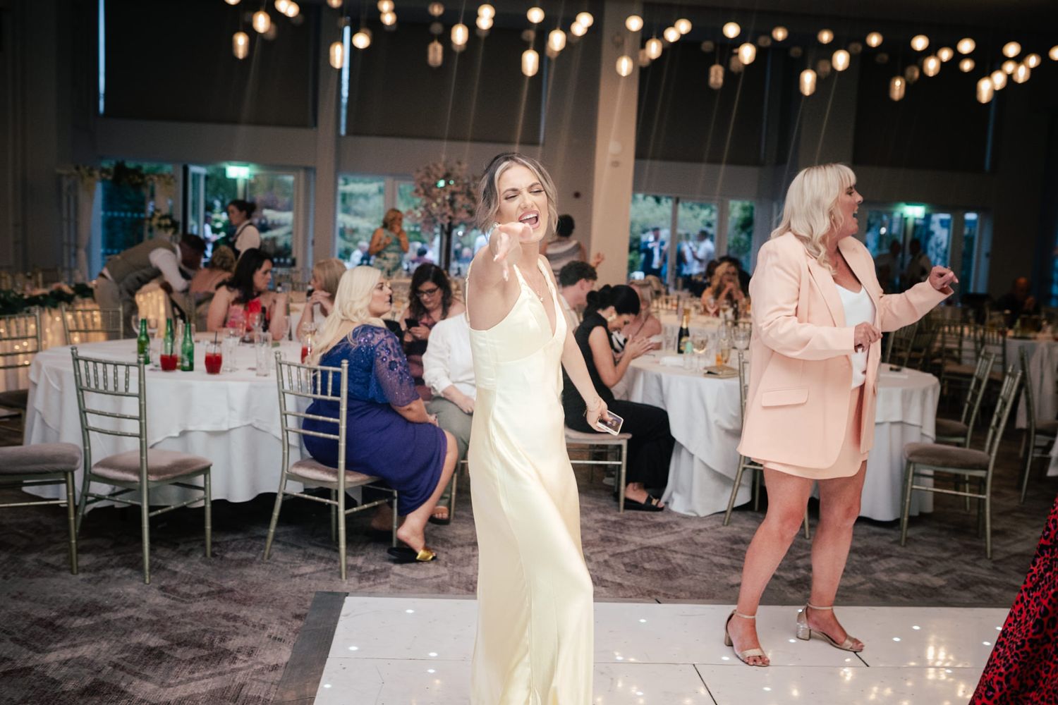 People dancing and celebrating at an elegant wedding reception with twinkling lights and white chiavari chairs in background.
