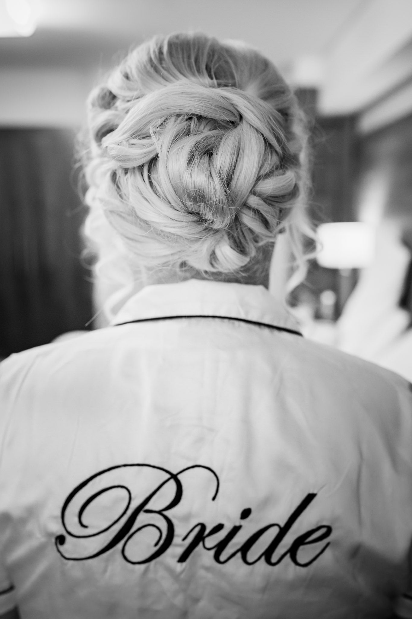 A black and white photo of an elegant bridal updo hairstyle and white robe with 'Bride' embroidered on the back.