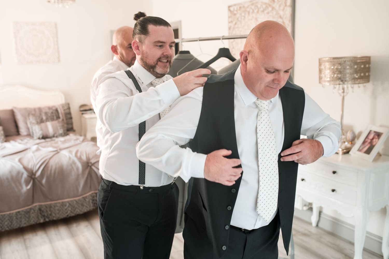 Two people in formal white shirts and black vests getting dressed for an event in a light-colored room.