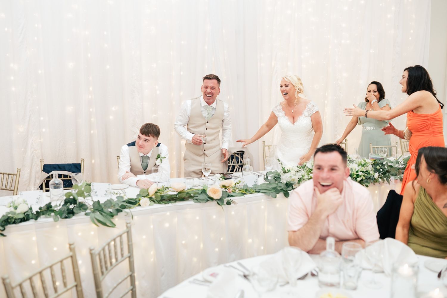 Wedding party and guests celebrate during reception in elegant white banquet hall with greenery decorations.