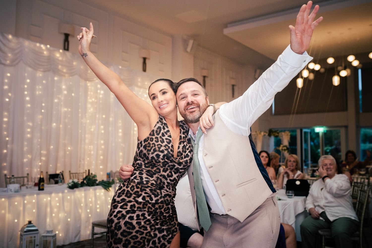 Two wedding guests dance joyfully together, raising their arms in celebration with string lights glowing in background.
