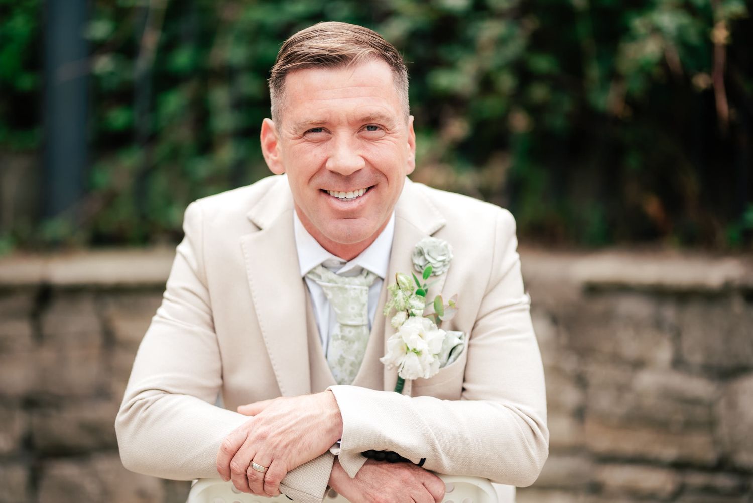 A person wearing a light beige suit jacket and boutonniere poses in an outdoor garden setting.