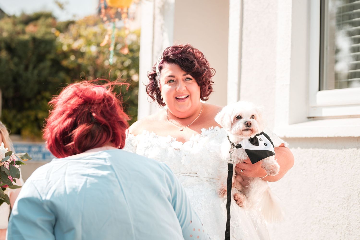 Smiling person in a white lace dress holds a small white dog while standing outside a house.