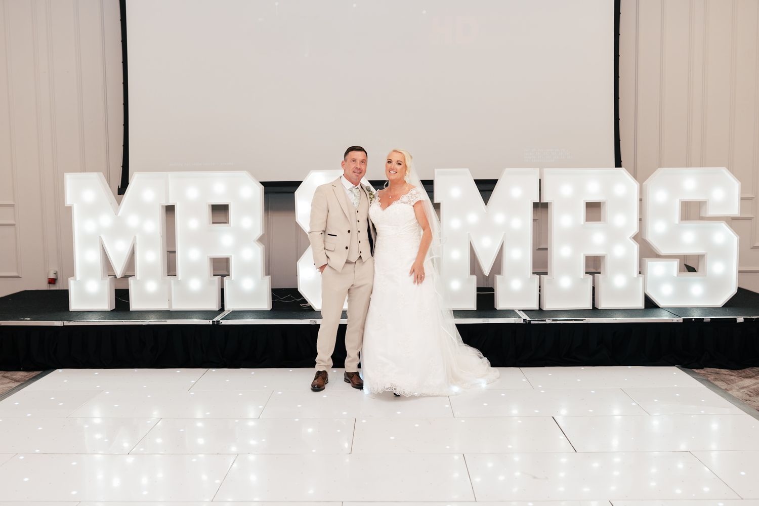 A couple poses in front of a large illuminated MR & MRS sign at their wedding reception.