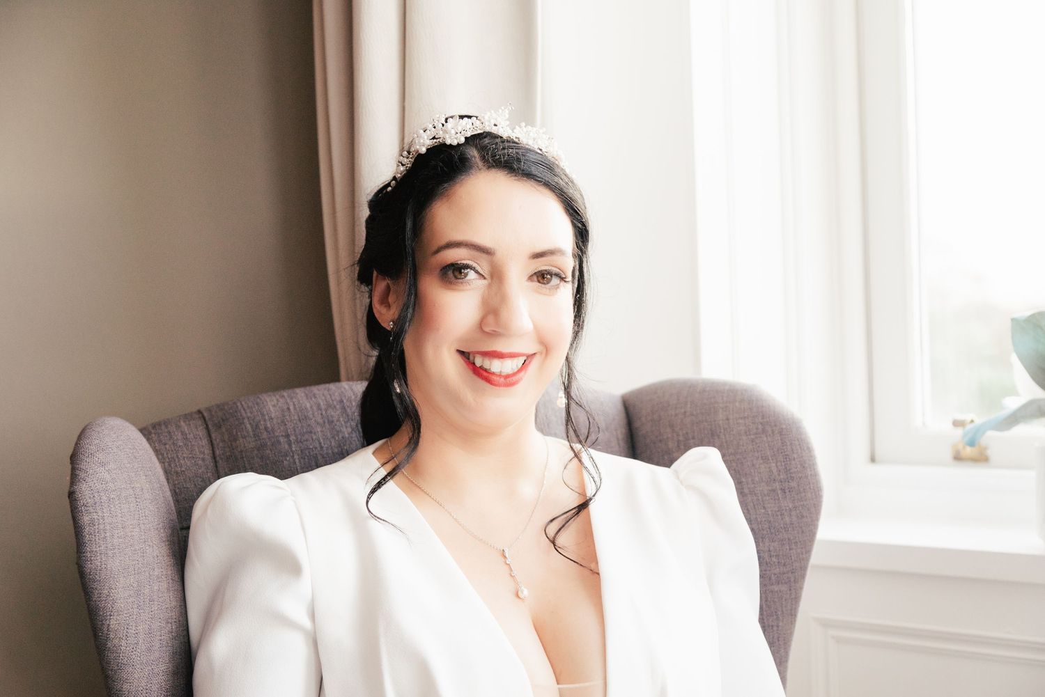 Elegant bride in white blazer smiling while seated in gray armchair by window.