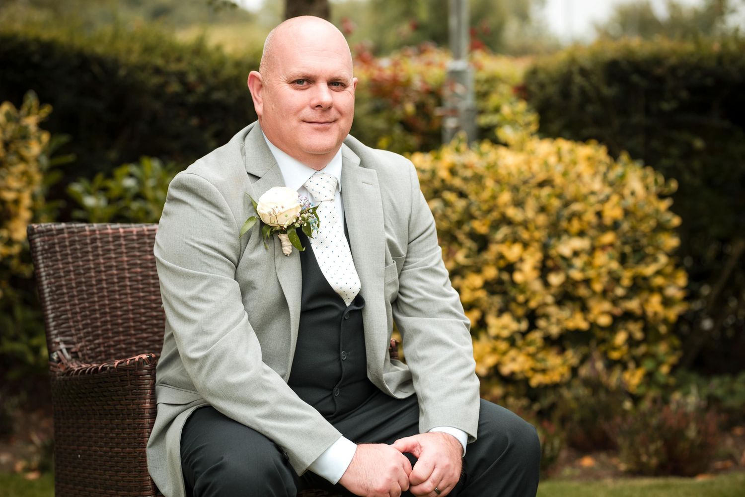 Wedding guest wearing light gray suit jacket and dark vest sits on wicker chair in garden setting with blooming shrubs.