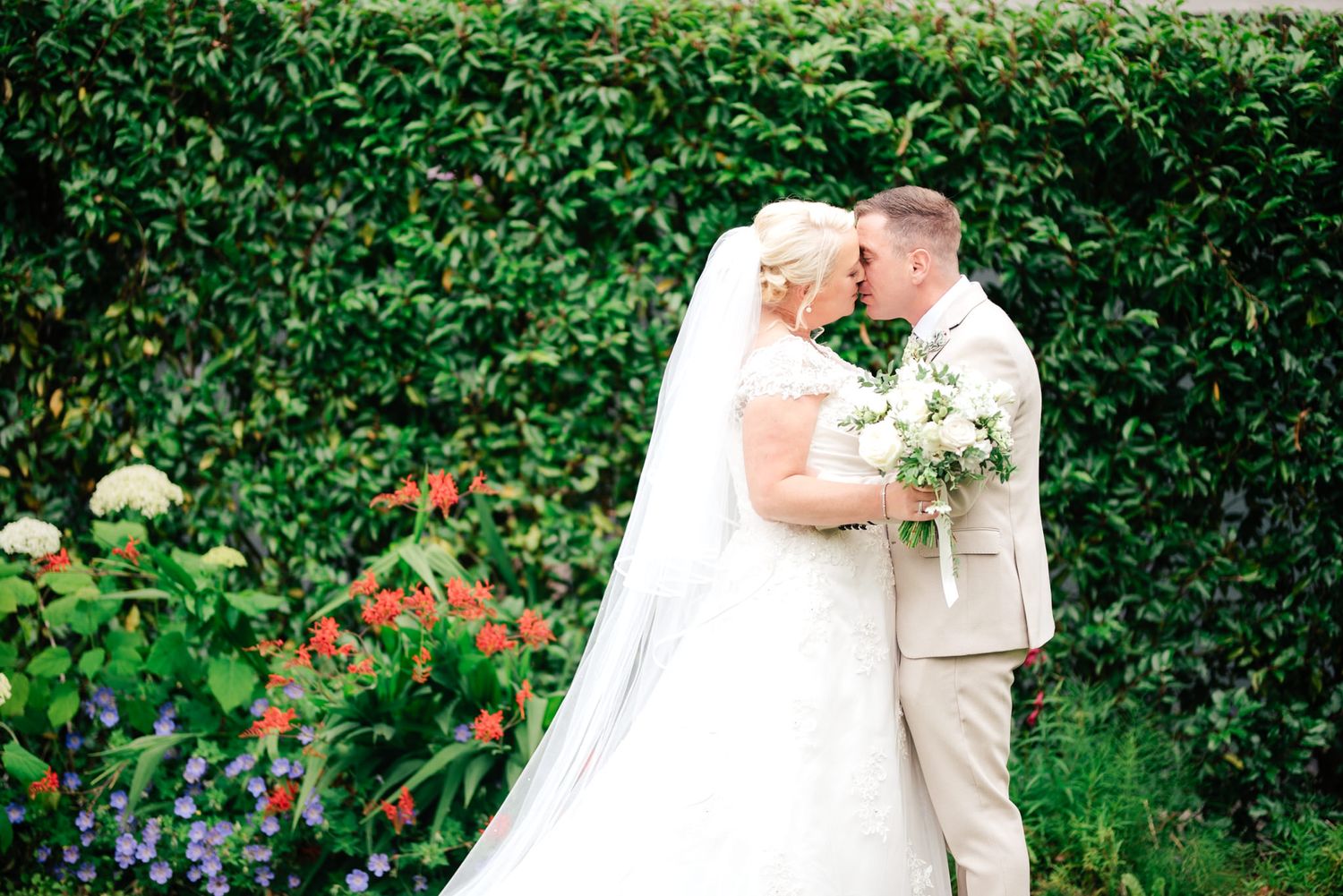 Beautiful wedding couple shares romantic embrace in garden setting surrounded by lush greenery and colorful flowers.