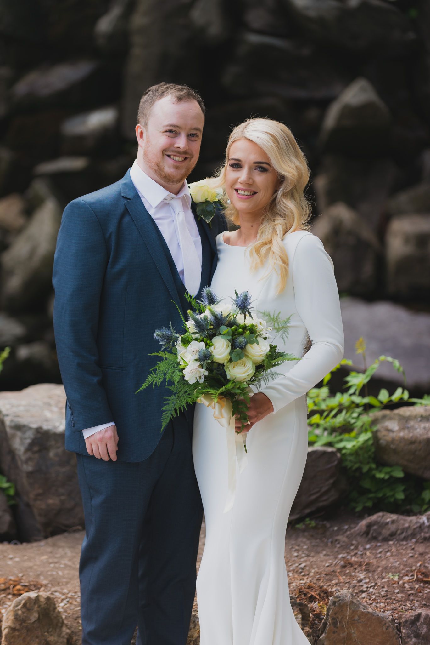 A happy couple posing for a wedding portrait against a natural stone backdrop.