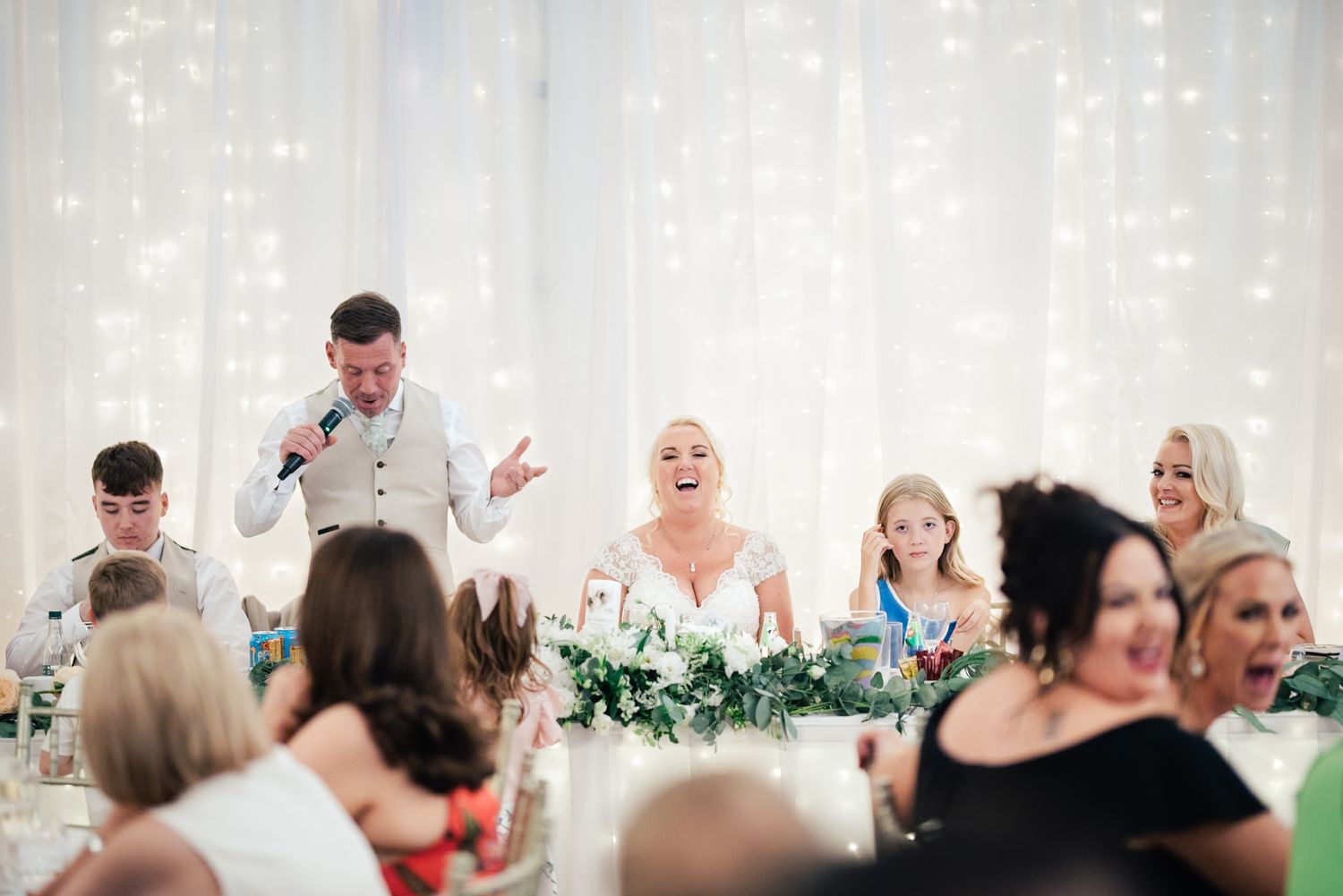 Wedding guests laugh and react during reception toasts at an elegant white-decorated indoor venue.