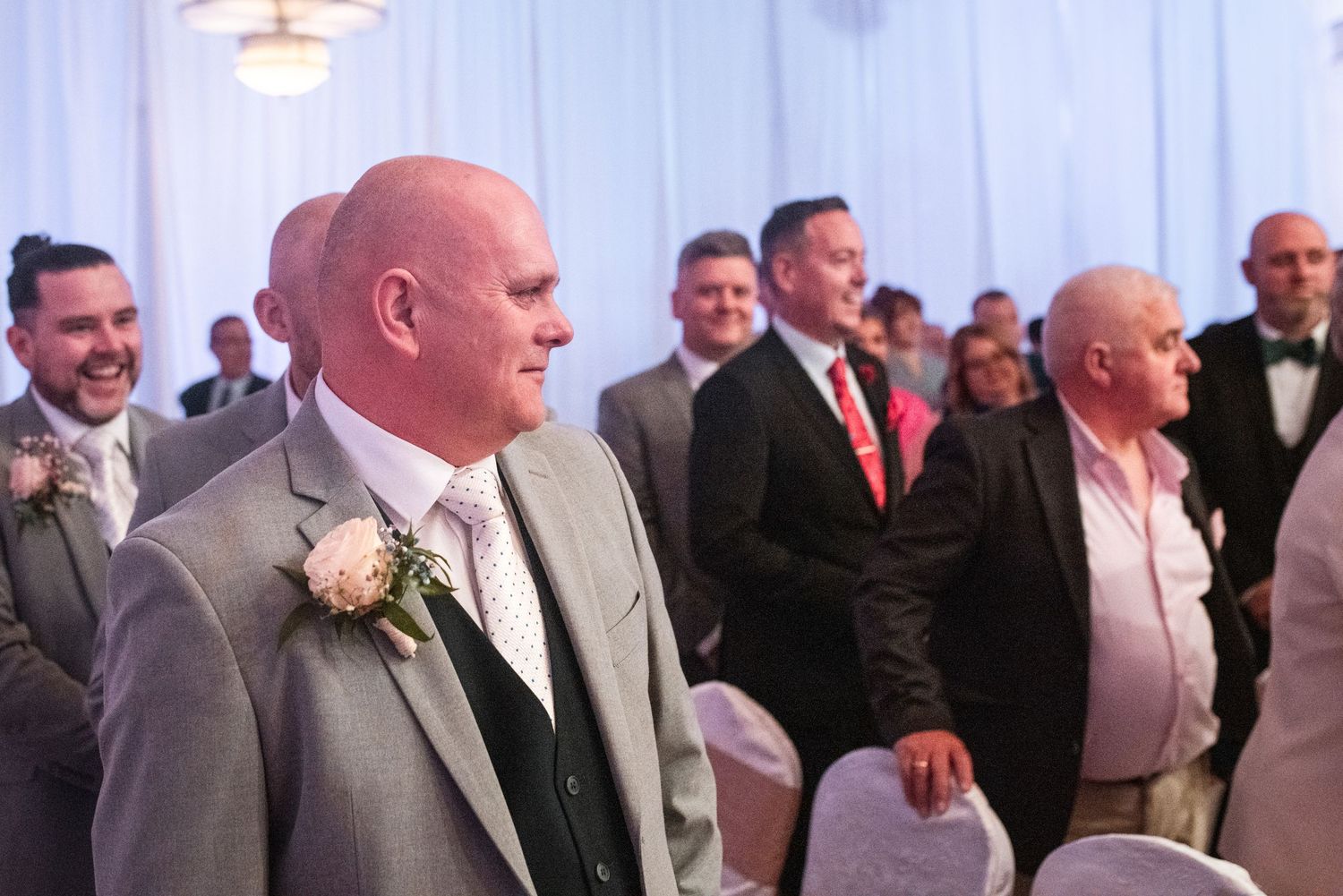 People in formal attire attend an elegant wedding ceremony in a white-draped indoor venue.