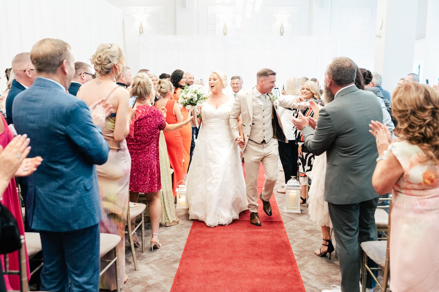 Wedding guests rise to their feet during a ceremony reception in an elegant venue with red carpet.