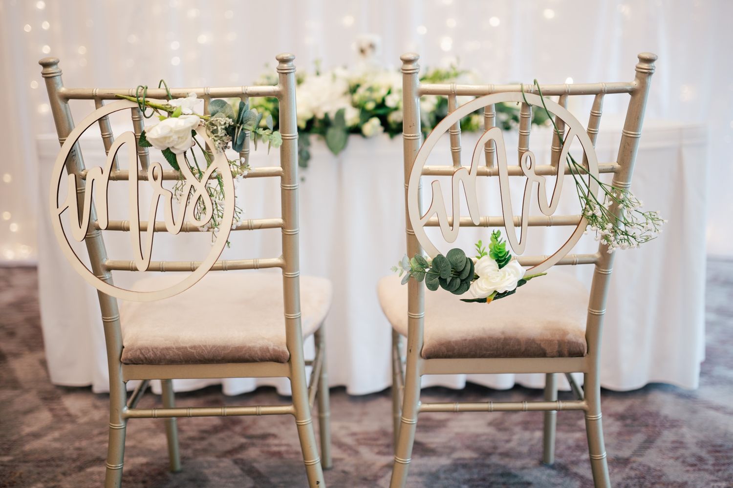 Two elegant wooden chairs adorned with 'Mr & Mrs' circular signs and greenery at a wedding reception setup.