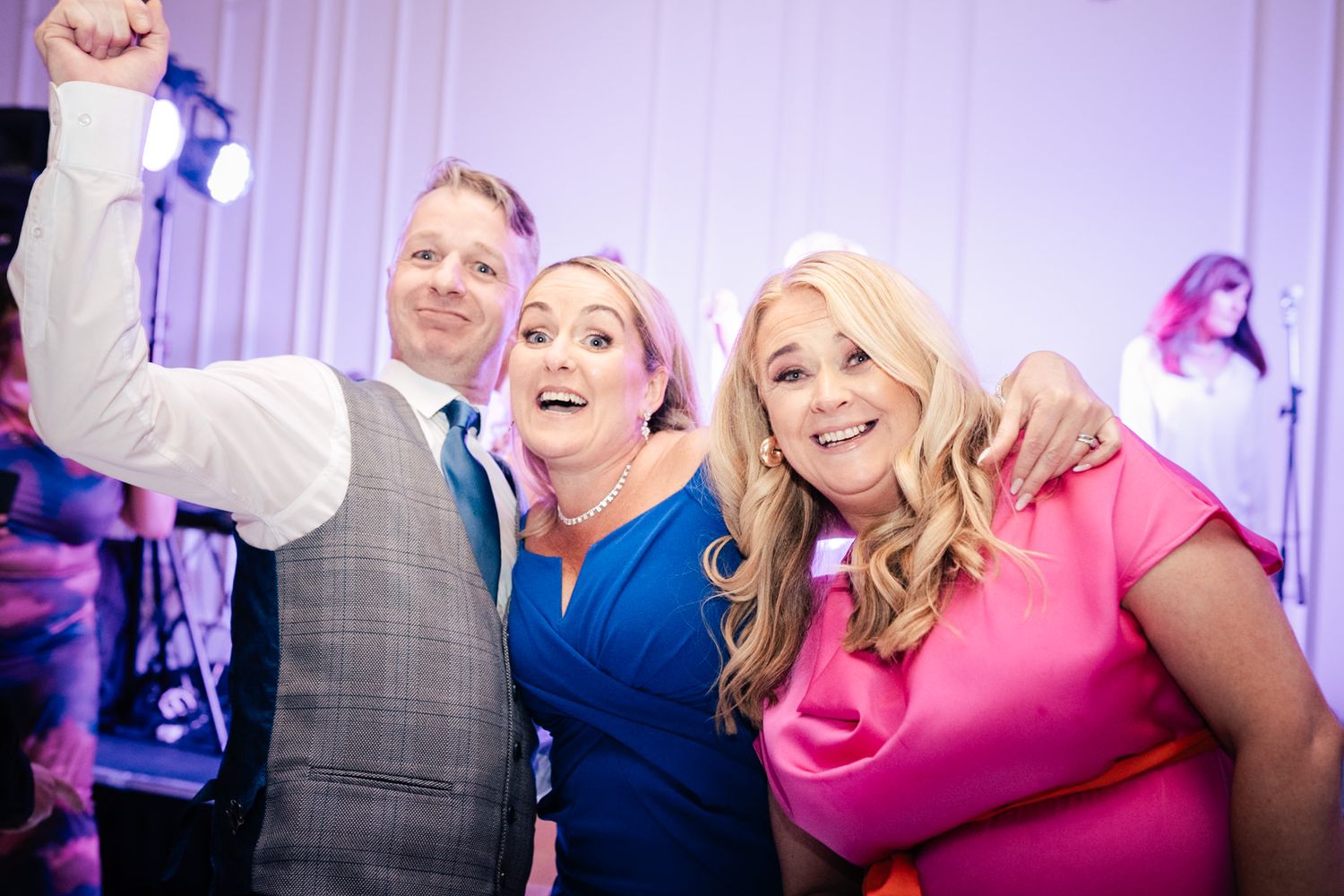 Three party guests having fun and posing together at a wedding reception with purple lighting in the background.