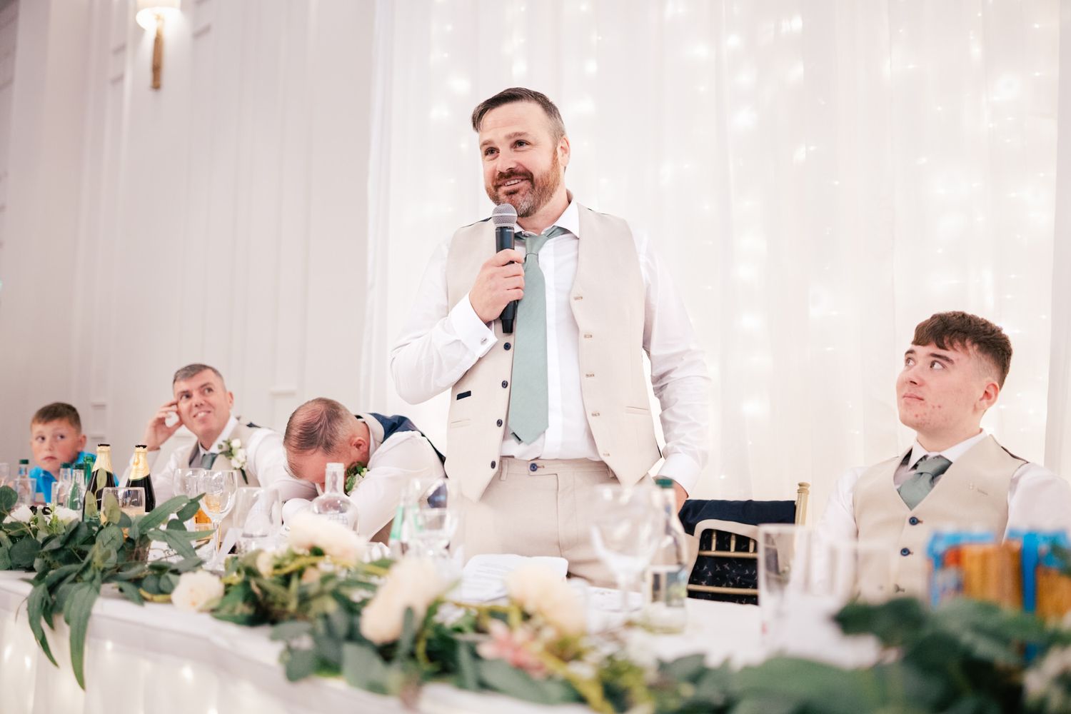 Speaker delivers wedding toast at elegant head table adorned with greenery garland and white decor.