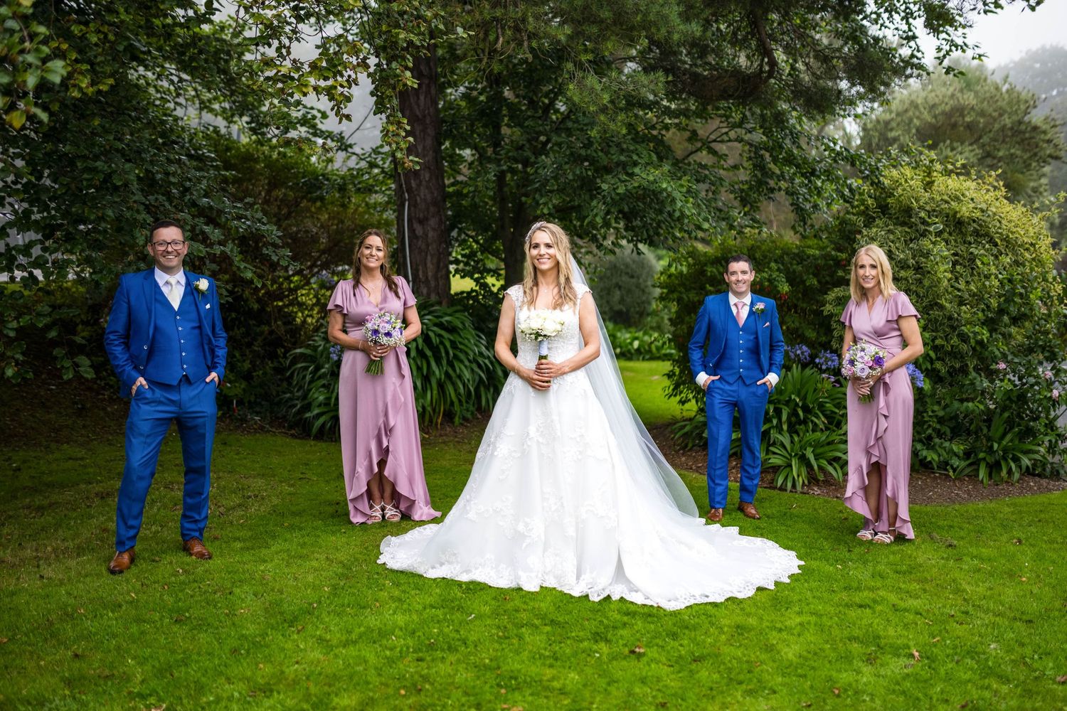 Wedding party in vibrant blue suits and pink dresses pose on a lush green lawn with trees in the background.