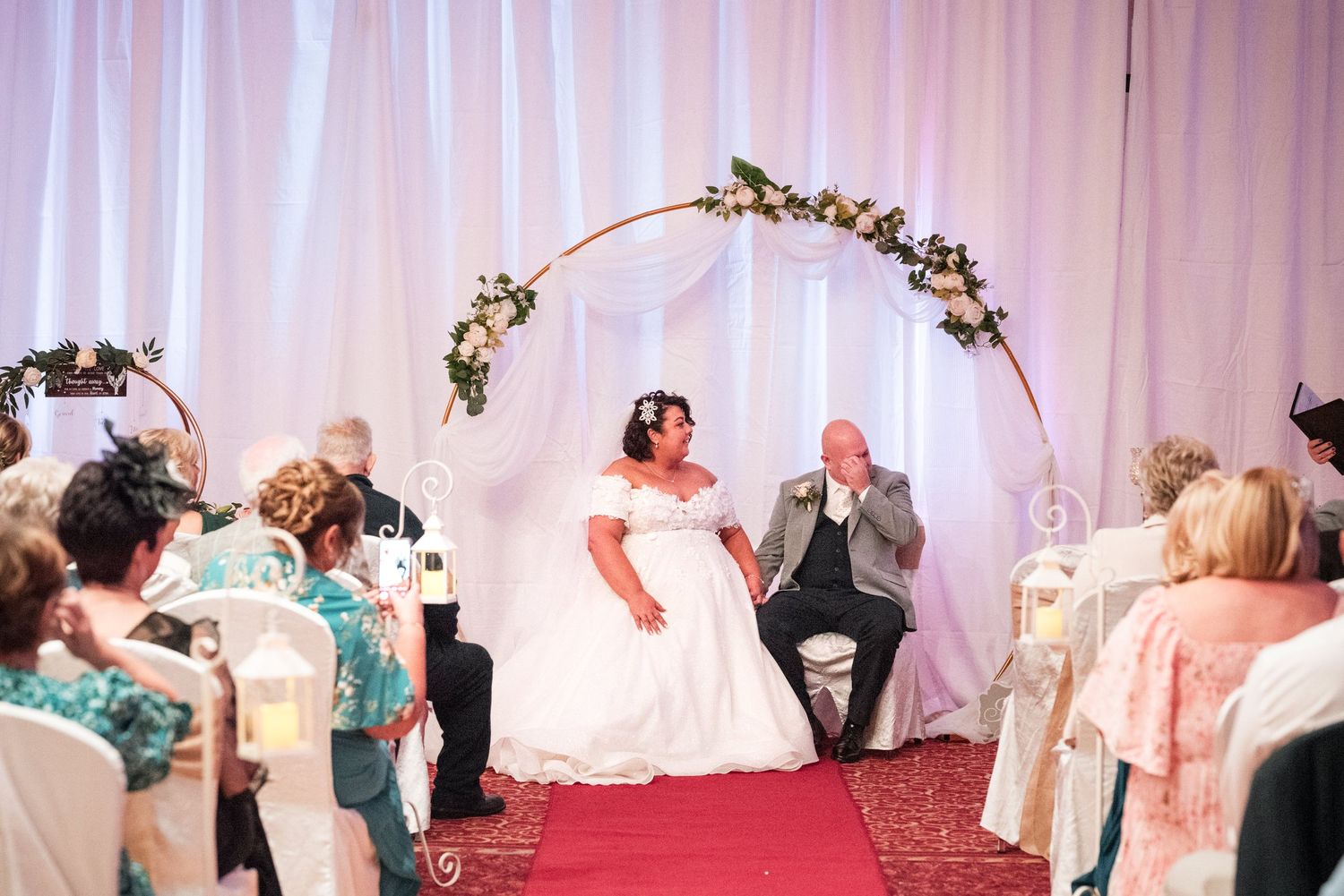 Wedding ceremony takes place under floral arch with couple seated on stage as guests watch from decorated chairs.