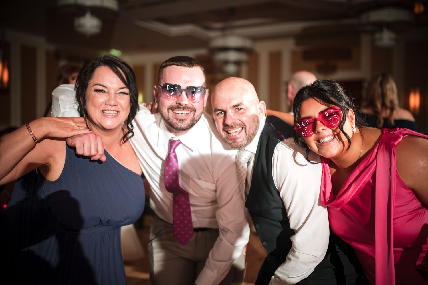 Four party guests posing together at a dimly lit event, with one wearing red heart-shaped party glasses.