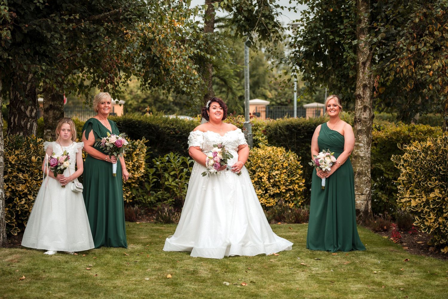 Four people in formal attire stand together in a garden setting with green hedges and trees in the background.