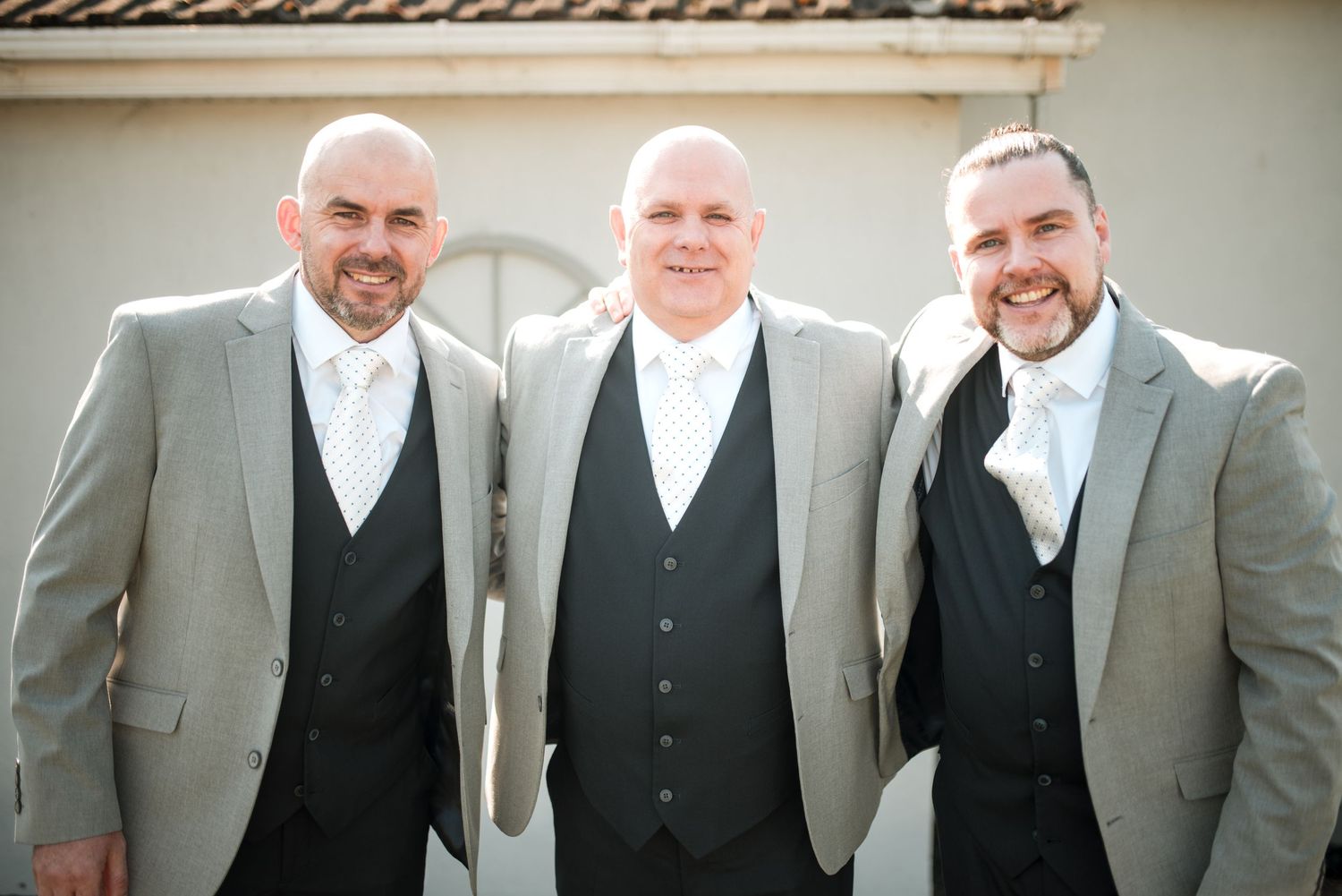 Three groomsmen wearing matching gray suits with black vests and white ties pose together smiling at a wedding.