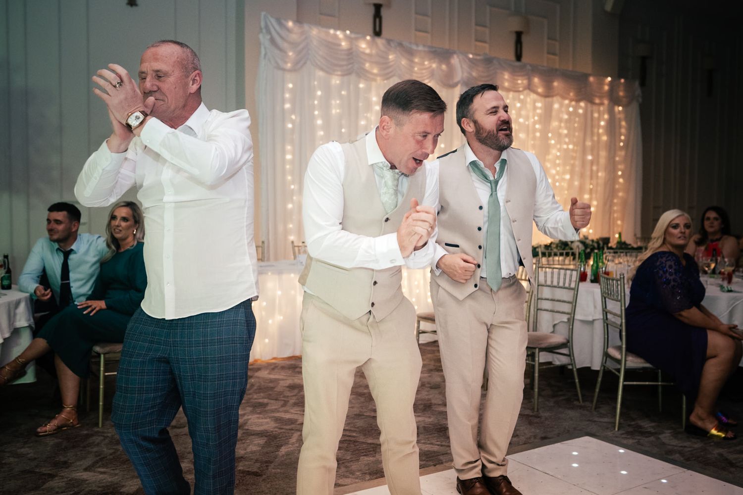 Dancing guests celebrate on the dance floor at a wedding reception with stringed lights in the background.