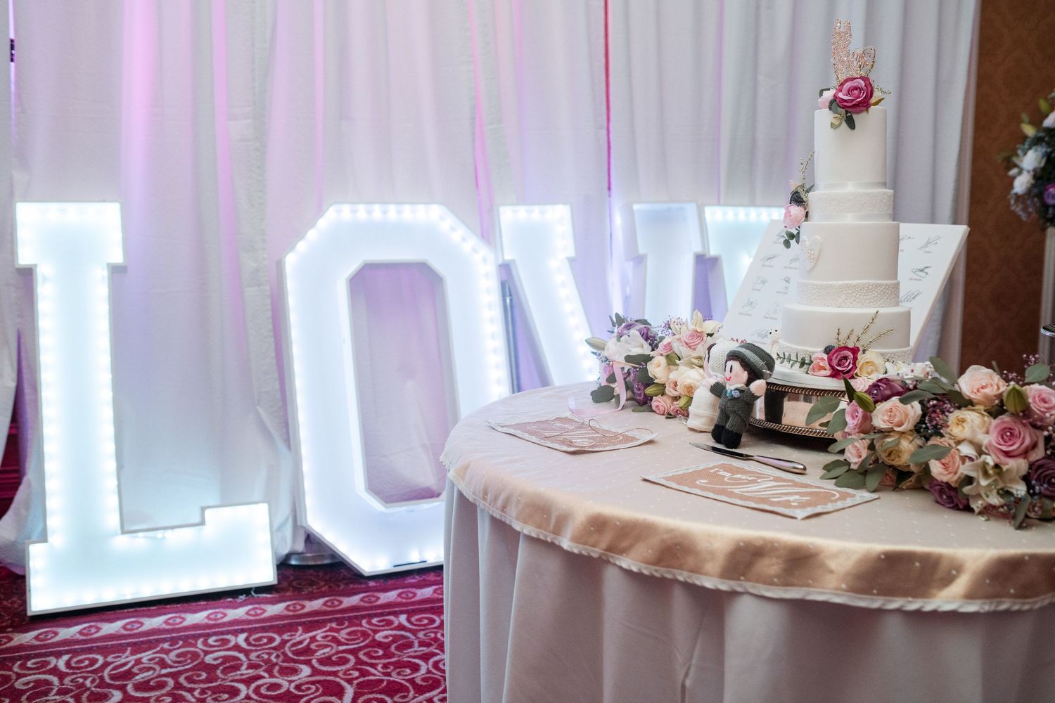 Elegant white wedding reception hall with pink lighting, sheer curtains and floral centerpieces on round tables.