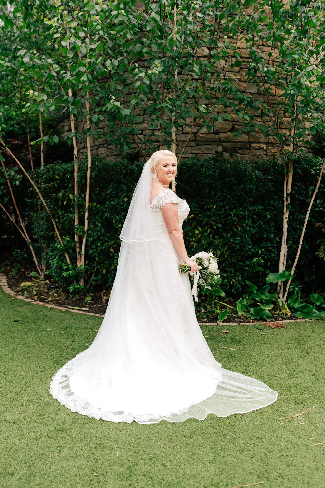 A bride wearing a flowing white lace wedding gown sits gracefully on emerald green grass against a wall of lush foliage.