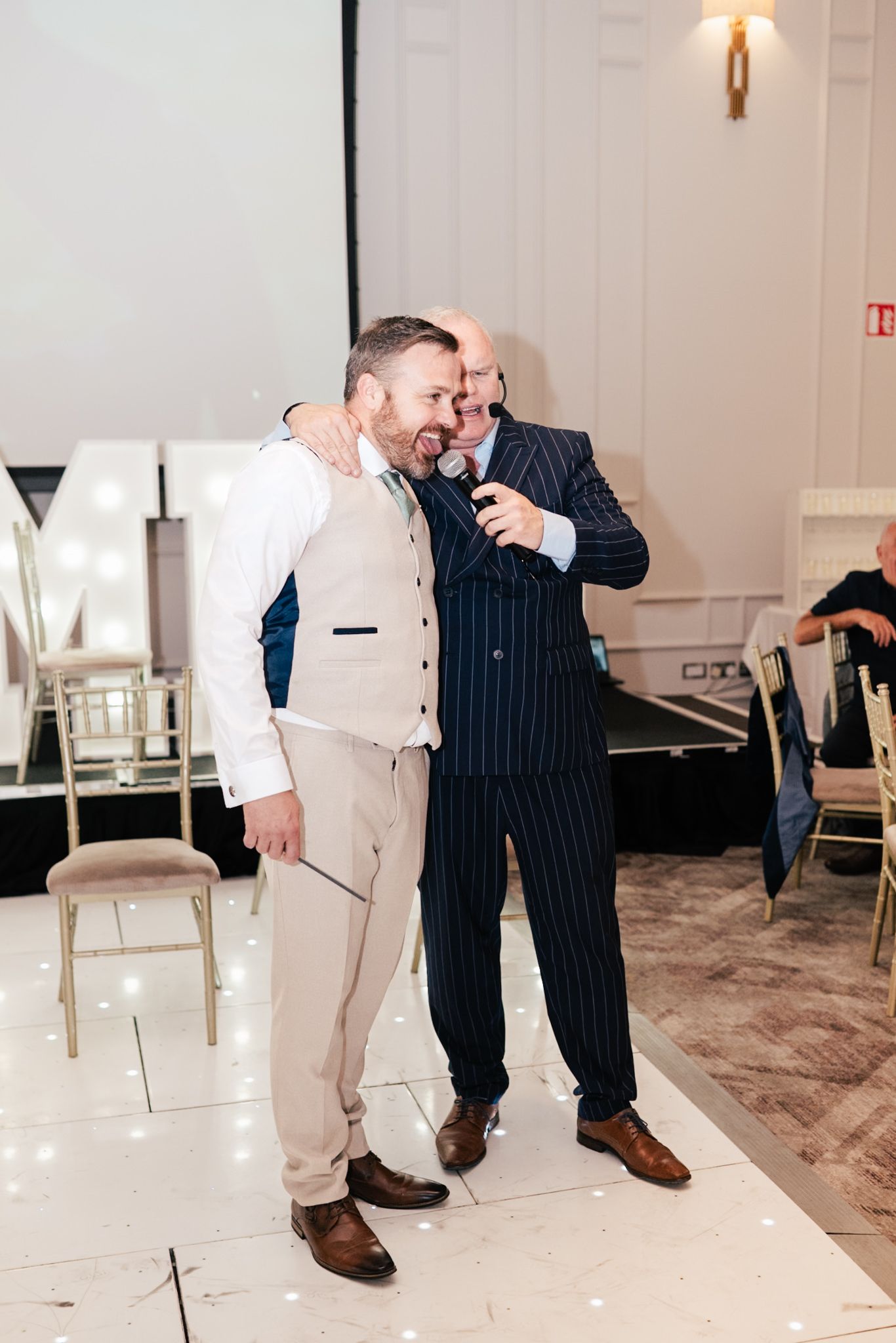 Two people share a lighthearted moment at a formal event, one in a pinstripe suit and one in beige attire.