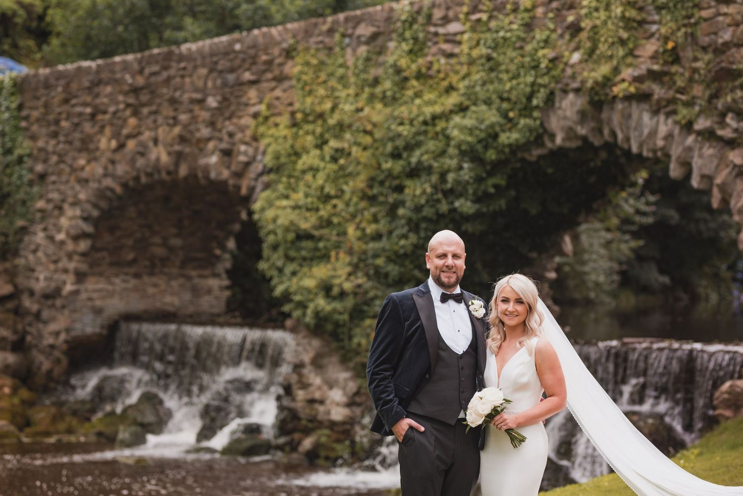 Wedding couple poses in front of scenic stone bridge and waterfall during autumn photoshoot.