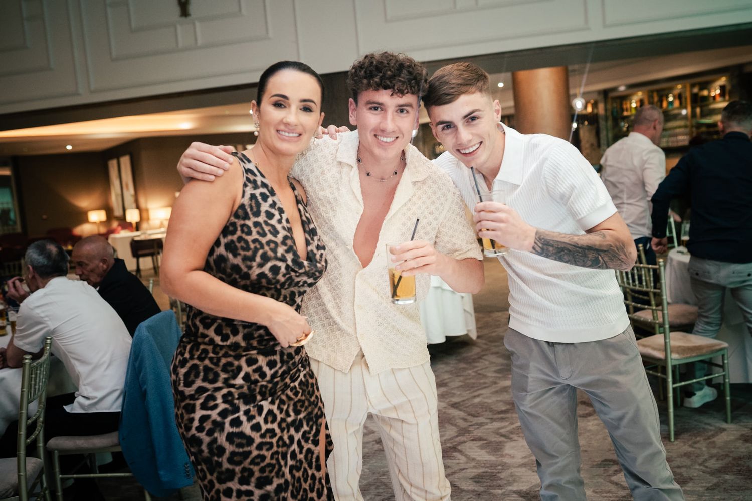 Three people smile together at what appears to be a restaurant or bar, with one wearing a leopard print dress.