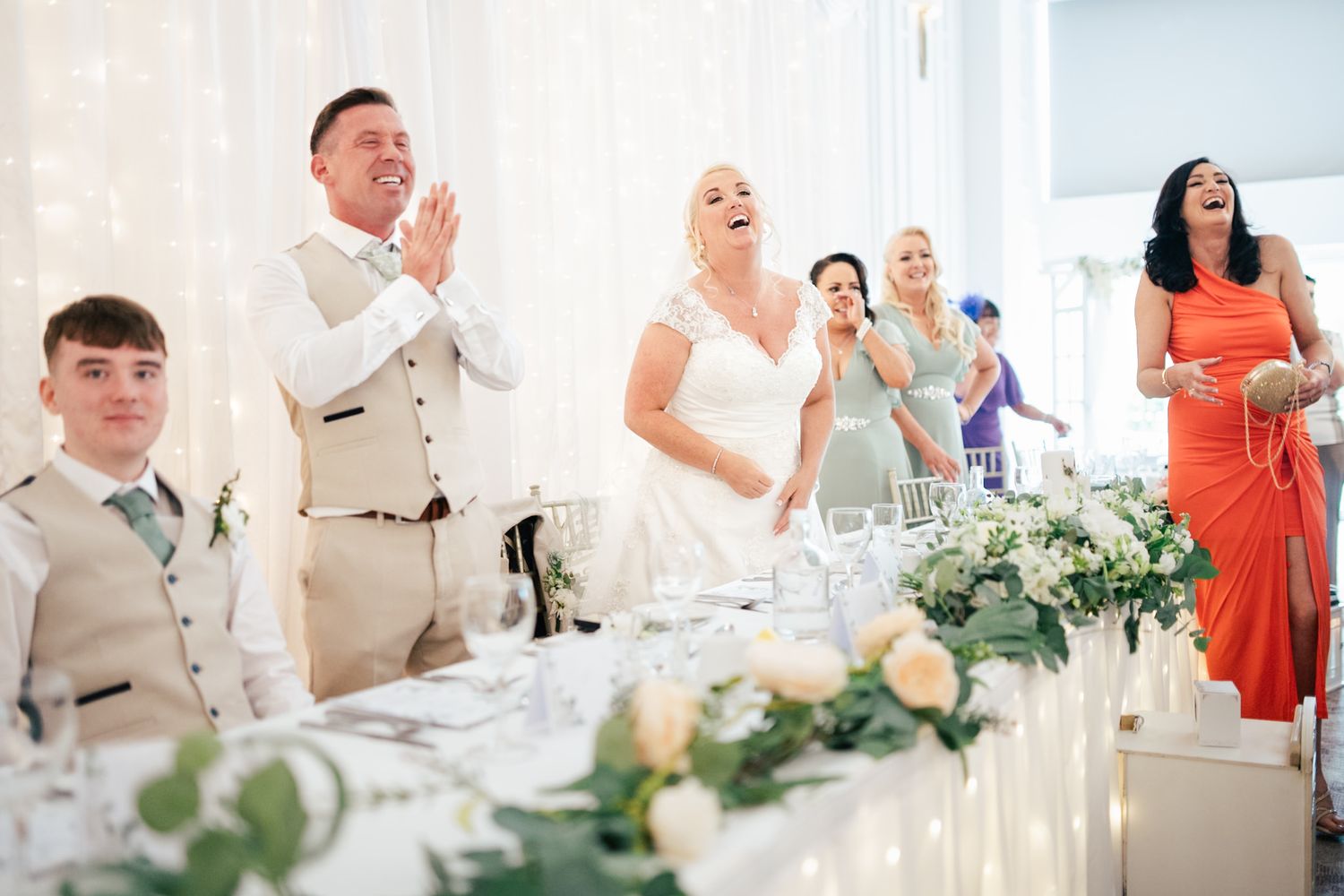 Wedding guests and newlyweds share a joyous moment at reception table decorated with elegant white floral arrangements.