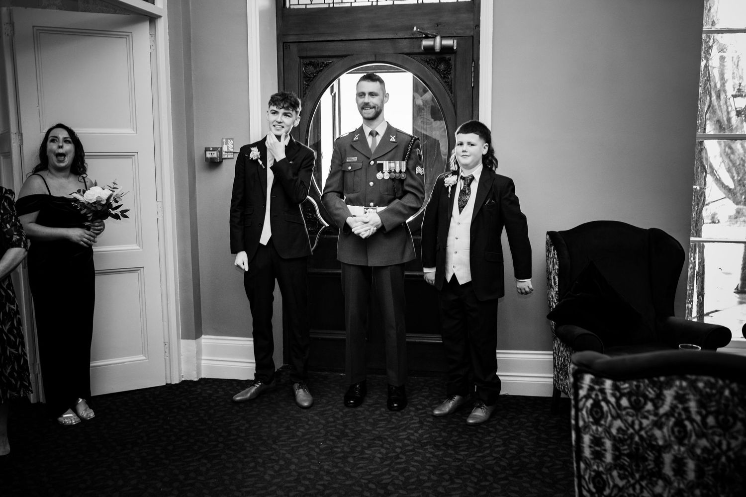 Black and white photo of a wedding party standing in elegant attire in a formal room interior.
