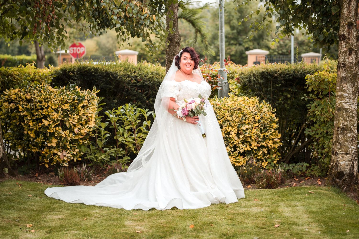 A bride in a white lace gown poses in a vibrant garden setting with blooming yellow flowers and greenery.