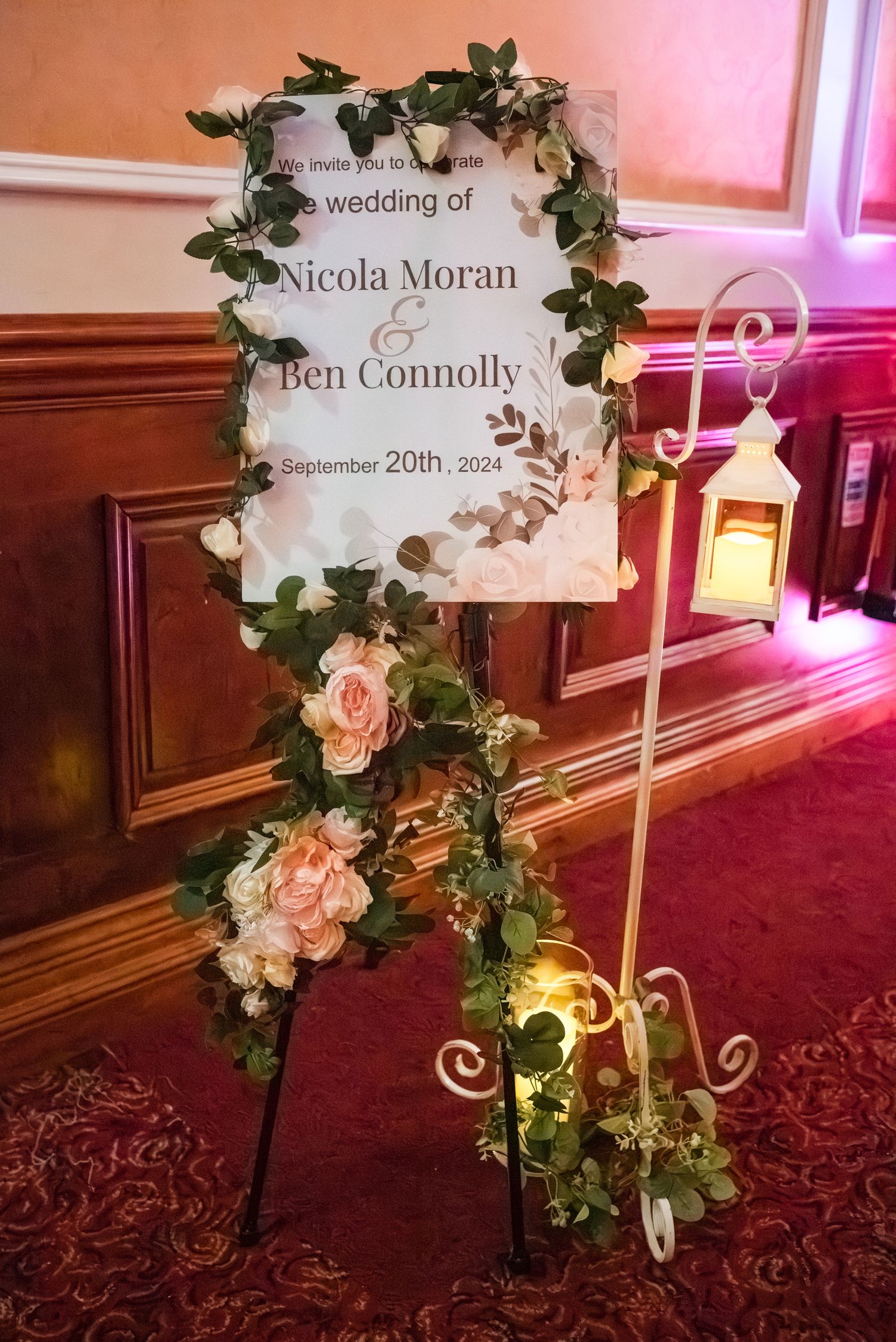 Elegant wedding seating sign featuring ivory flowers and greenery decorations on a gold stand against a pink-lit wall.