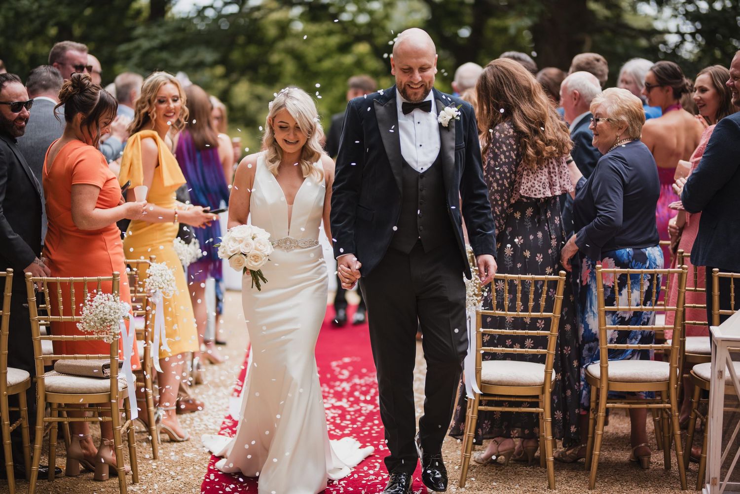 A couple walks down a rose petal-covered aisle at an outdoor wedding ceremony with guests looking on from wooden chairs.