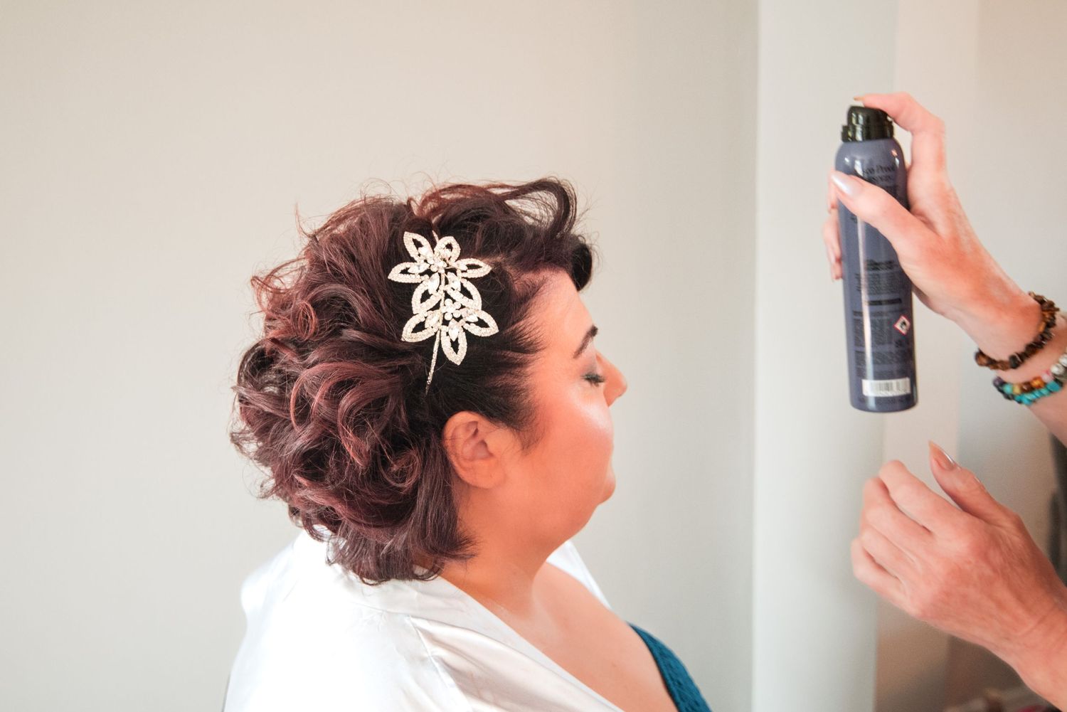 A stylist sprays hairspray on an elegant updo hairstyle adorned with a decorative hair accessory.