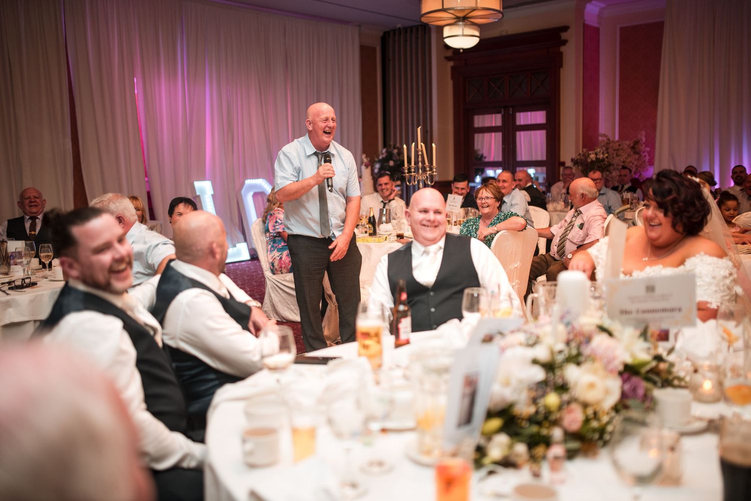 People enjoying laughter during wedding reception dinner as speaker gives toast at elegant venue with pink lighting.