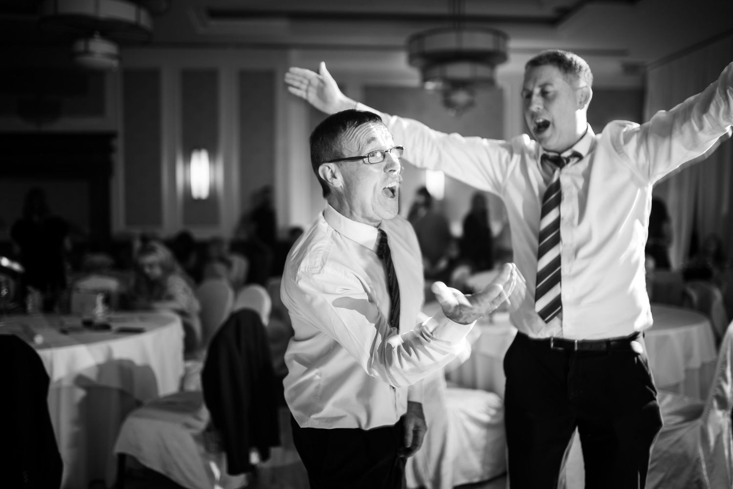 Two people in dress shirts and ties joyfully dance together at a reception venue in black and white photography.