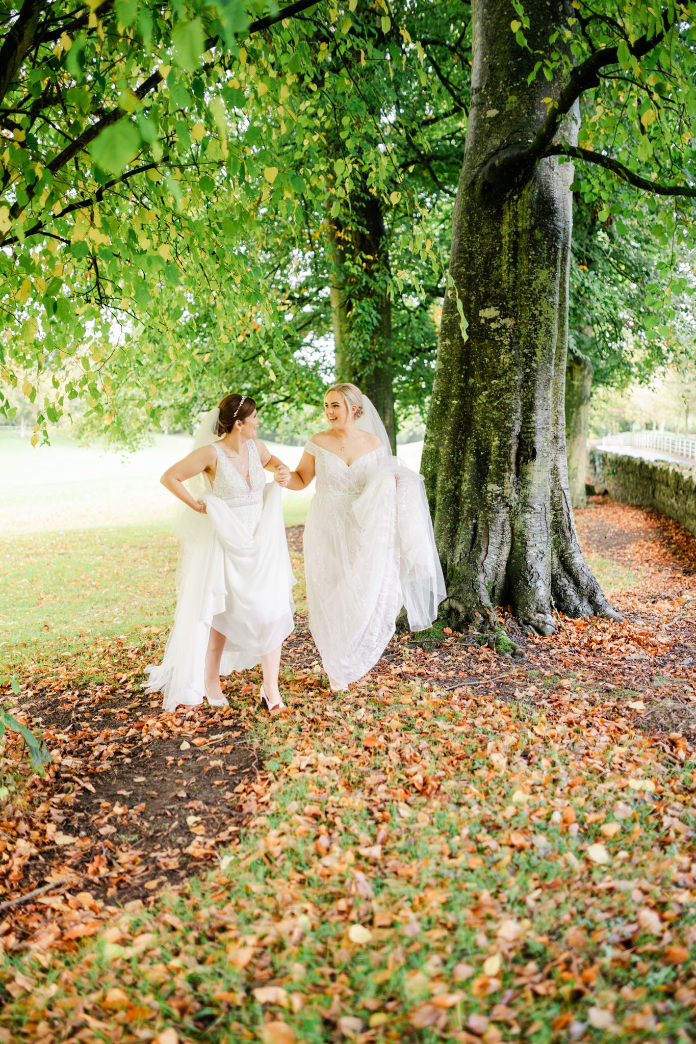 Two people in flowing white dresses share a playful moment under a large tree with fallen autumn leaves.