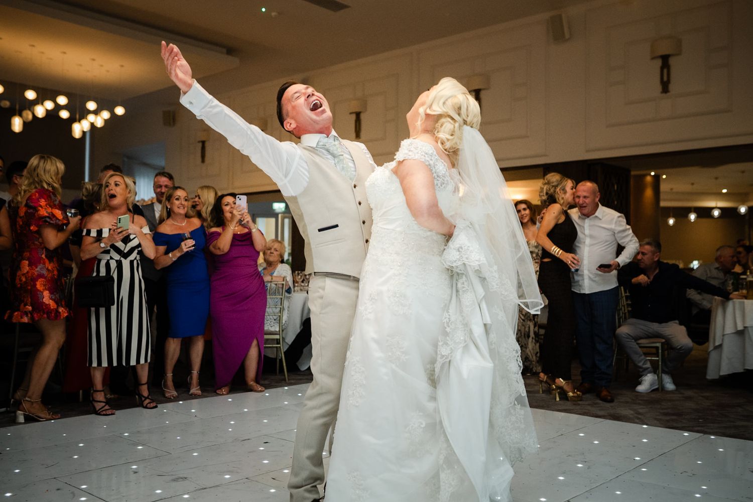 Two newlyweds dance joyfully on a white dance floor at their wedding reception while guests watch in the background.