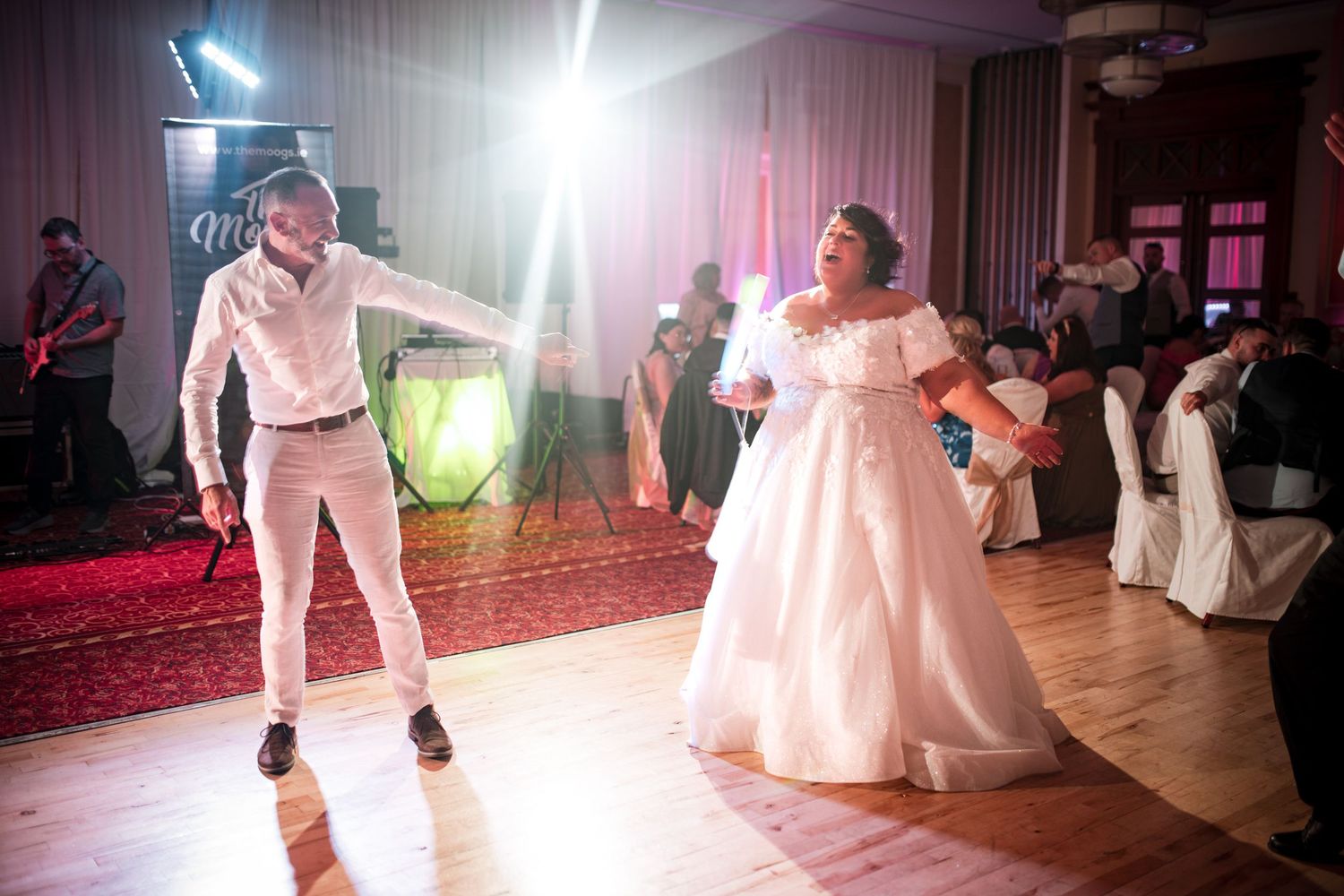 Two wedding guests dance energetically on an illuminated dance floor with pink lighting and decorative elements.