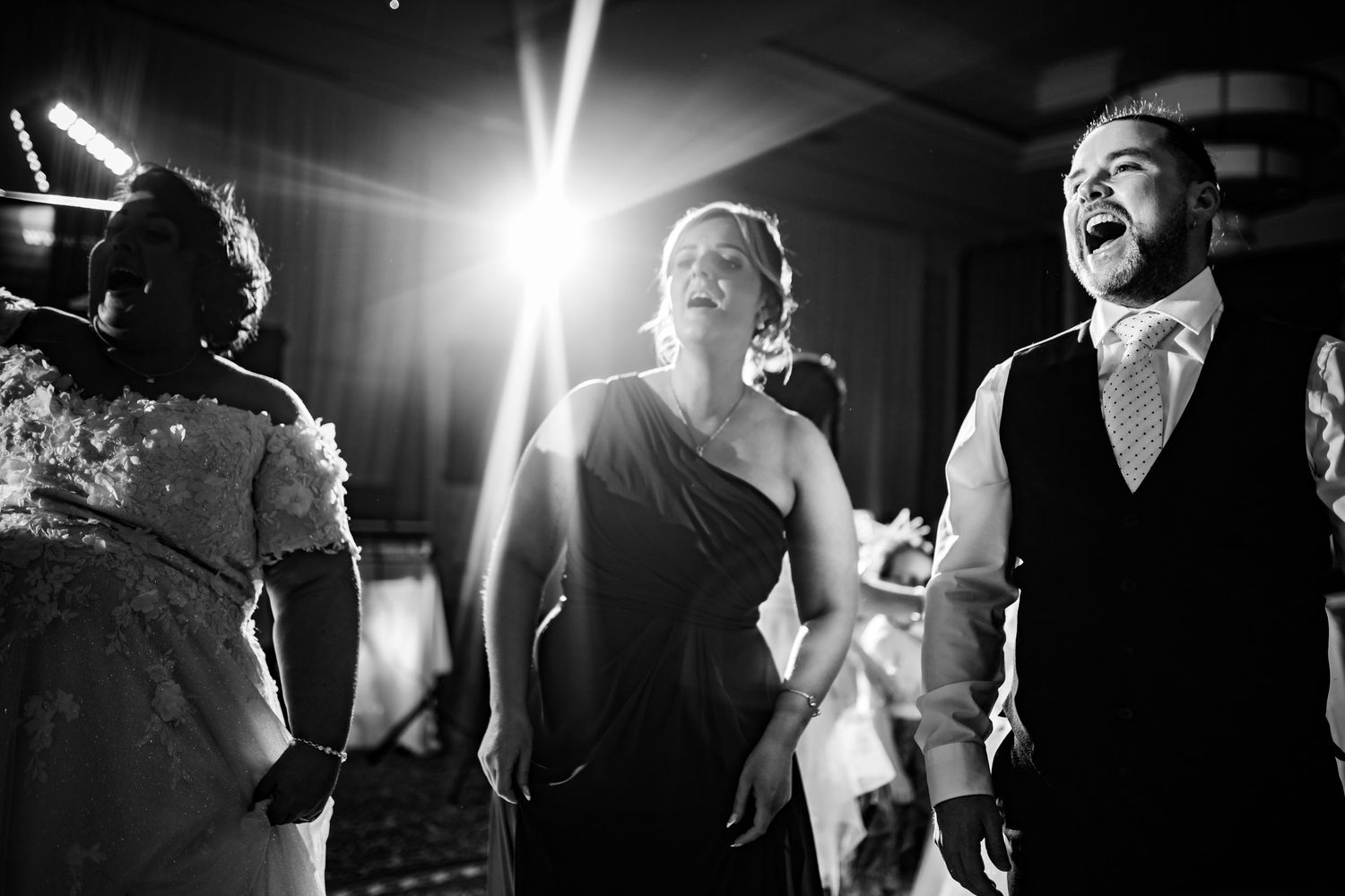 Wedding guests dancing energetically on a dark dance floor illuminated by bright spotlight beams during reception celebration.