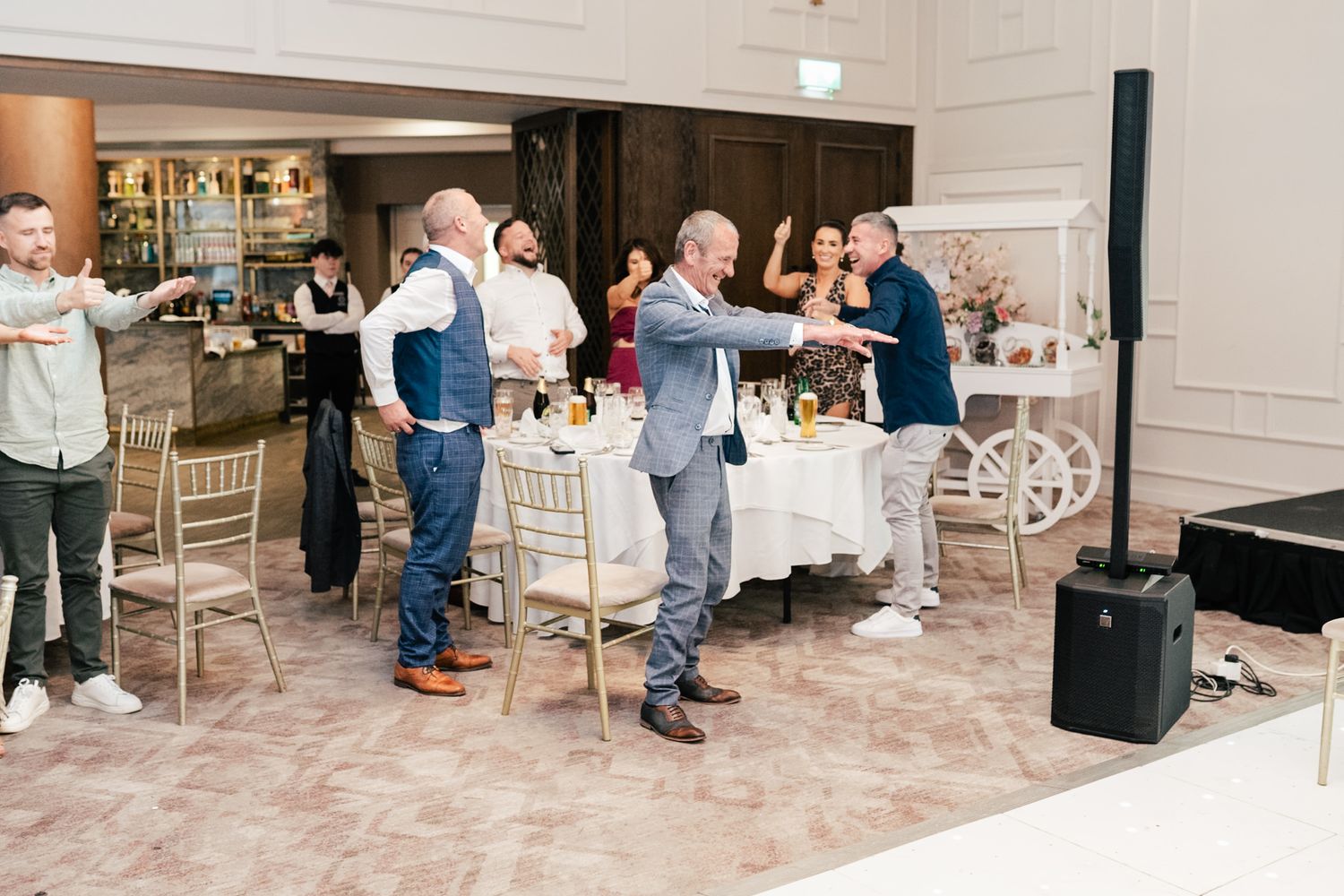 A panoramic view of people gathered in a white-walled venue space during a social event, with tables and chairs arranged.