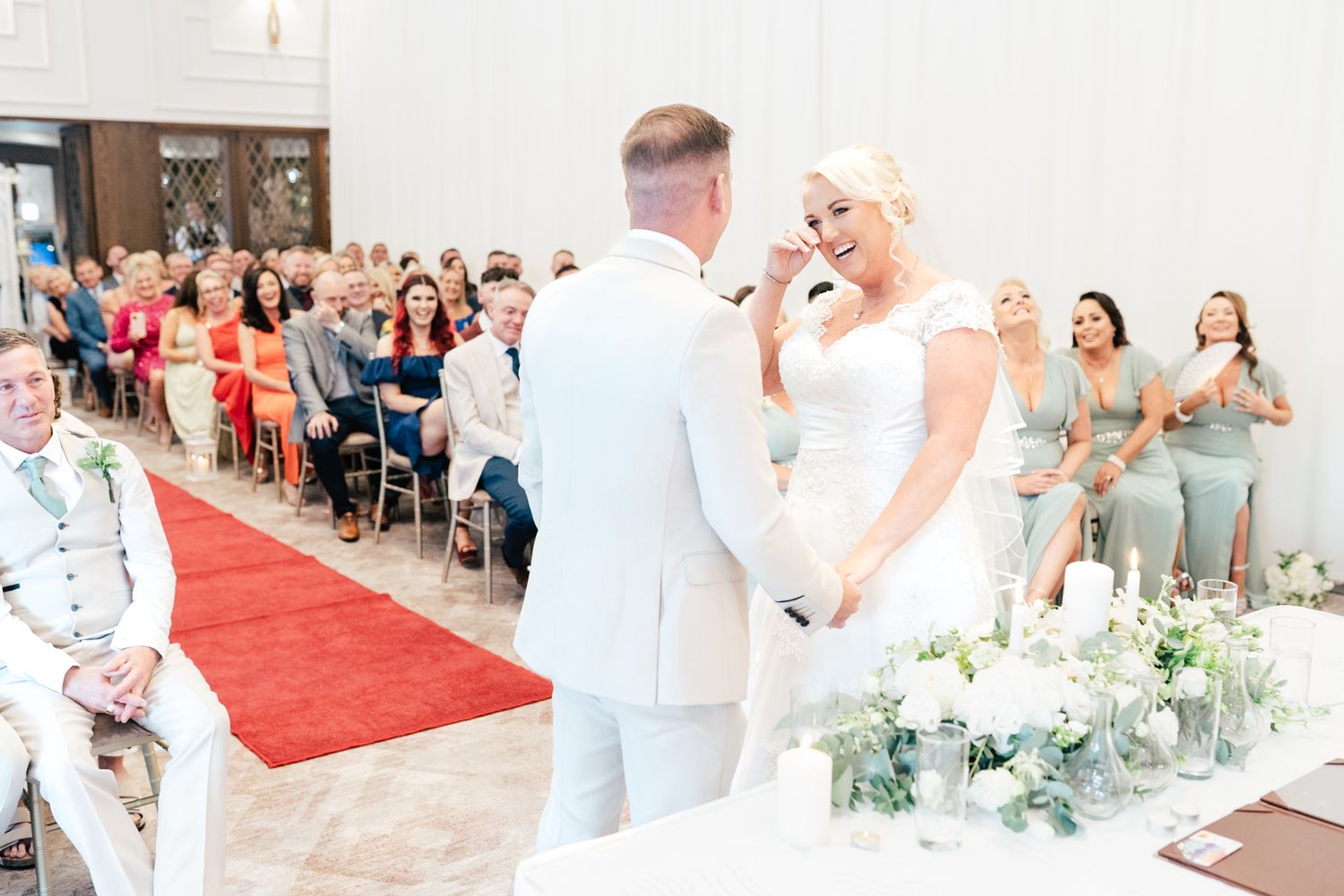 A bride and groom share a joyful moment during their wedding ceremony with attendants in sage green dresses looking on.
