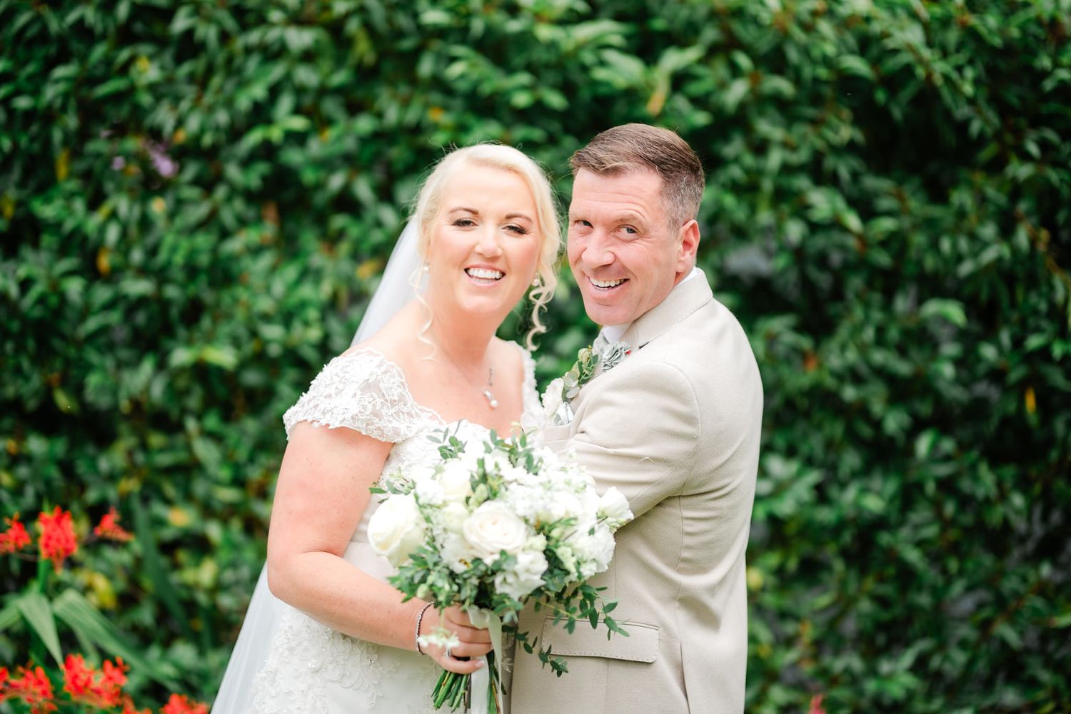 A couple laughs and embraces in their formal wedding attire in front of a lush green garden backdrop.