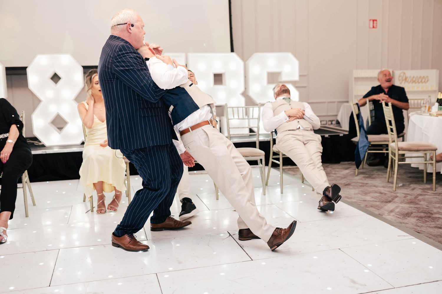 Two people performing an energetic dance move during a wedding reception with guests watching.